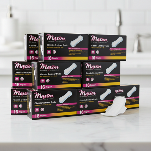 Stack of Maxim Hygiene ‘Classic Contour Pads’ boxes (16 Regular) arranged on a bathroom countertop, with one white pad displayed in the foreground.