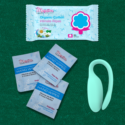 Flat lay on a textured green surface: a large pack of ‘Maxim’ organic cotton intimate wipes at the top, three small ‘Maxim’ antibacterial disinfecting hand wipes sachets fanned out below on the left, and a mint-green curved device on the right.