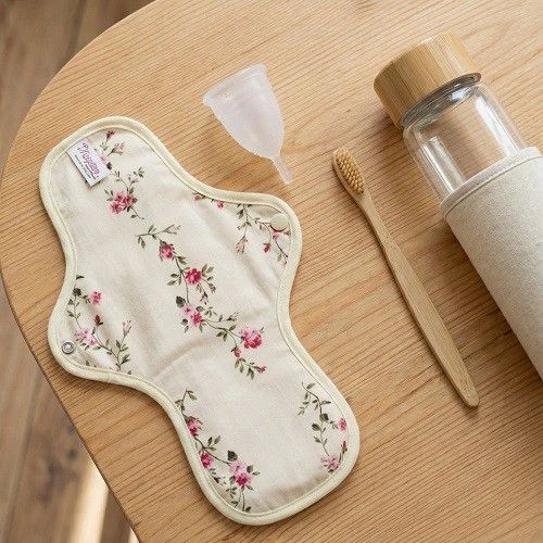 Reusable floral cloth menstrual pad on a wooden surface with menstrual cup, bamboo toothbrush, and reusable bottle, highlighting sustainable zero-waste period care essentials.