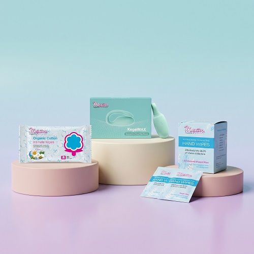 Flat lay on a textured green surface: a large pack of ‘Maxim’ organic cotton intimate wipes at the top, three small ‘Maxim’ antibacterial disinfecting hand wipes sachets fanned out below on the left, and a mint-green curved device on the right.